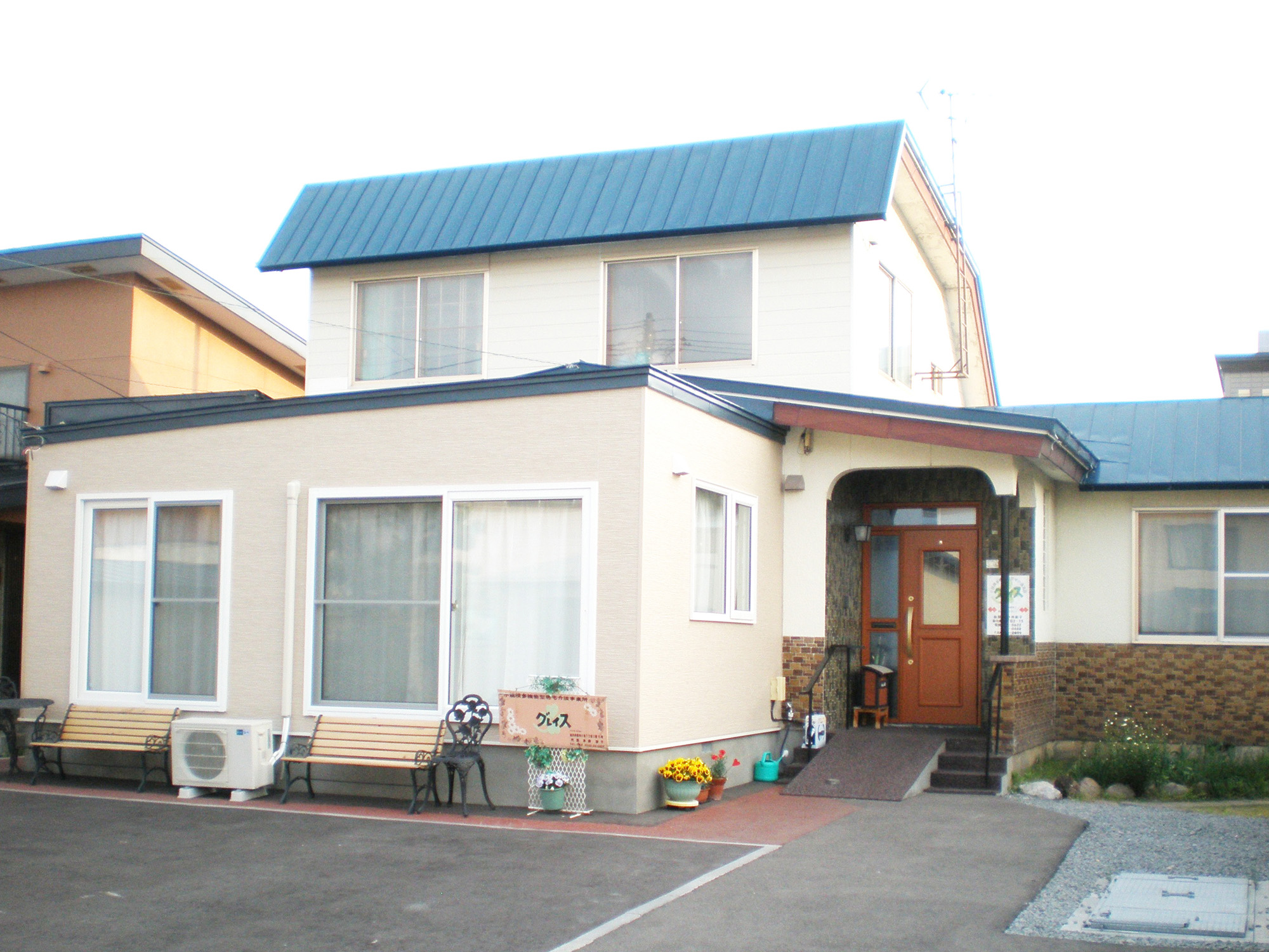青い屋根の介護施設の建物外観。玄関前に花や看板が配置